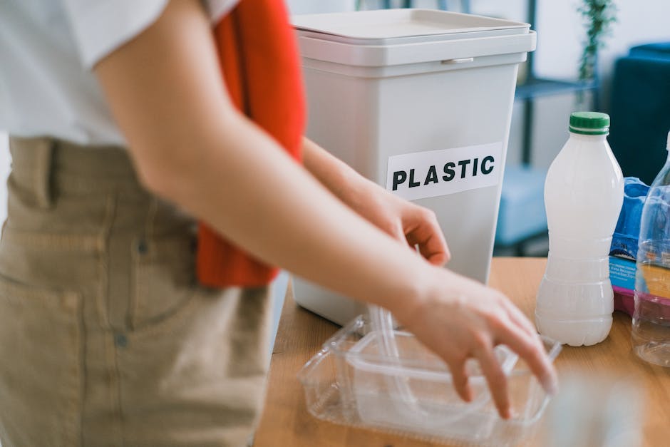 A young woman with long brown hair, dressed in a white t-shirt and a red sweater draped over her shoulders, stands in a modern kitchen holding a transparent plastic water bottle in her left hand and a