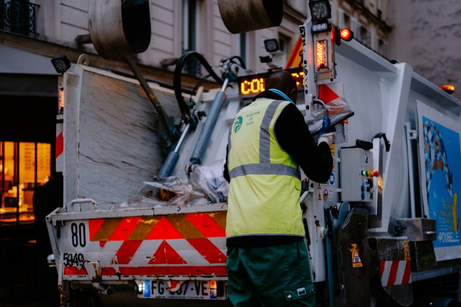 A waste collection operative wearing a high-visibility yellow vest with grey reflective stripes and dark clothing is positioned behind a large white rubbish truck, working on the vehicle's control pan