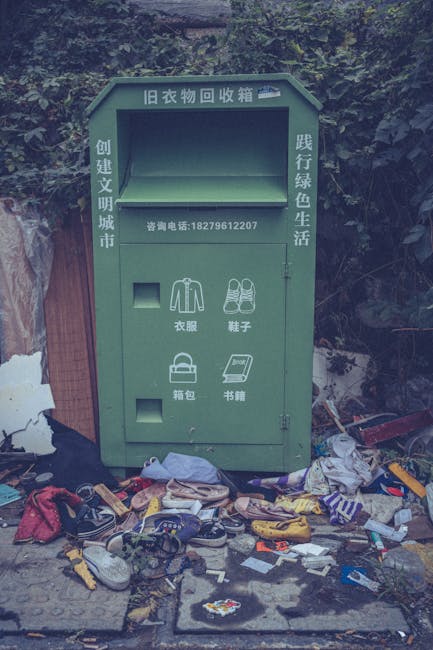 A large green clothing donation bin with Chinese characters and contact information printed on it, situated outdoors against a background of trees and foliage. The bin has multiple compartments for di